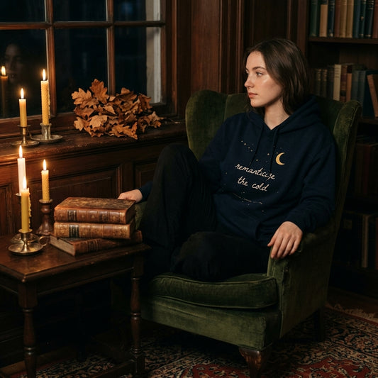 Woman wearing the navy Romanticise the Cold hoodie in a candlelit dark academia reading nook